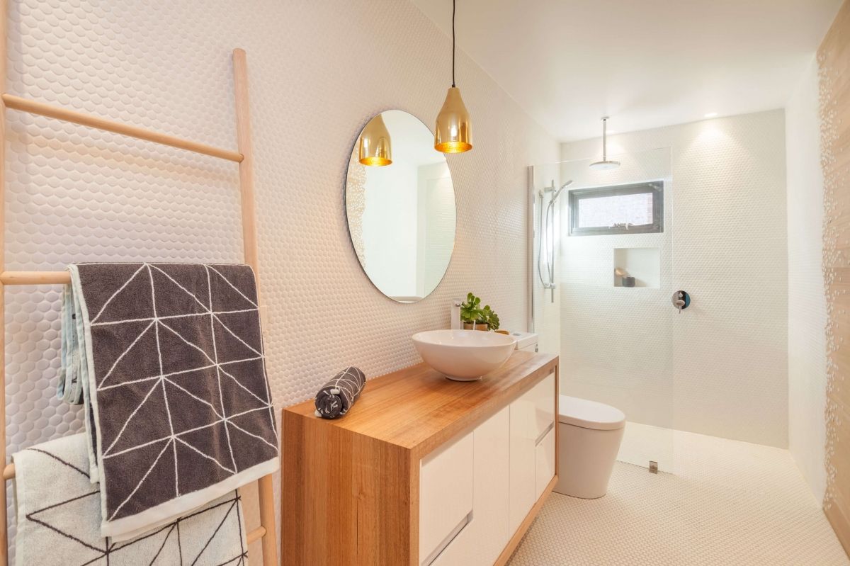 Bright bathroom vanity with wood cabinet, round mirror, pendant lights, and shower area in the background