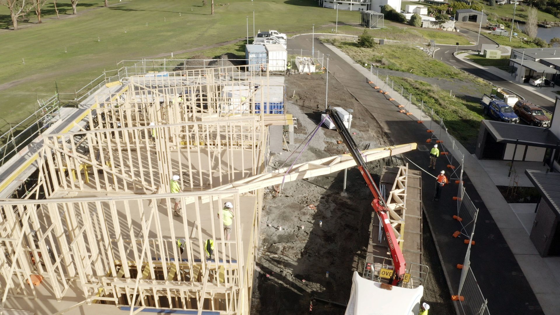 Construction site with wooden house framing and a crane beside a road on grassy land