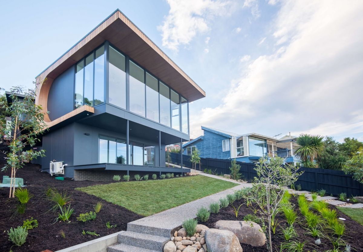 Modern two-story house with large windows, sloped roof, and landscaped front yard on a hillside.