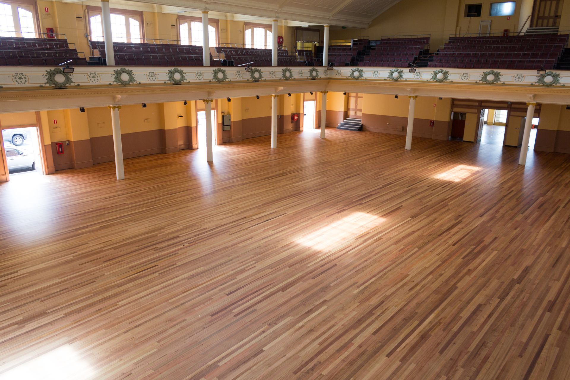 Empty wooden gymnasium with polished floor and upper-level balcony seating, lit by sunlight