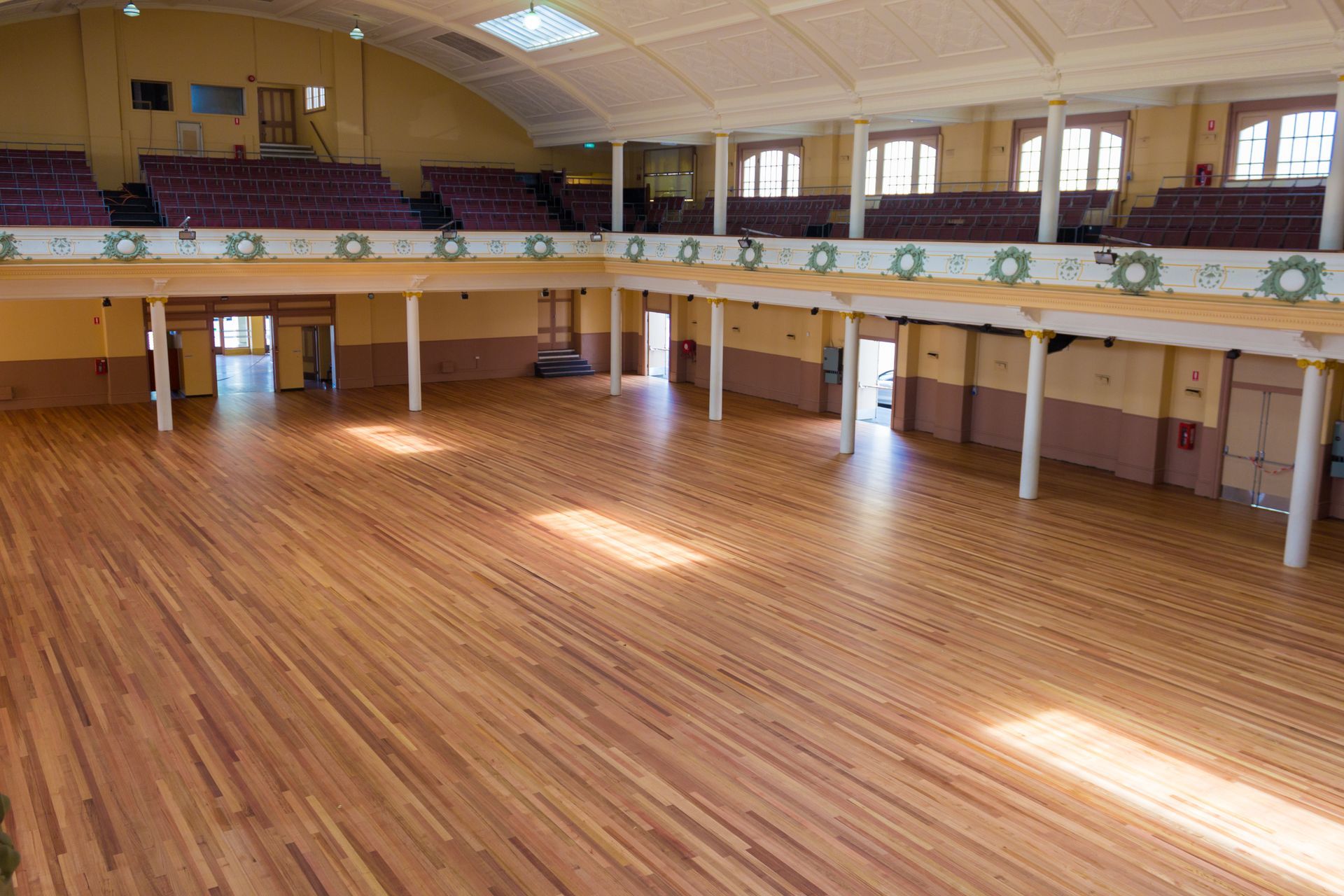 Empty indoor gymnasium with polished wooden floor, balcony seating, and bright overhead lights