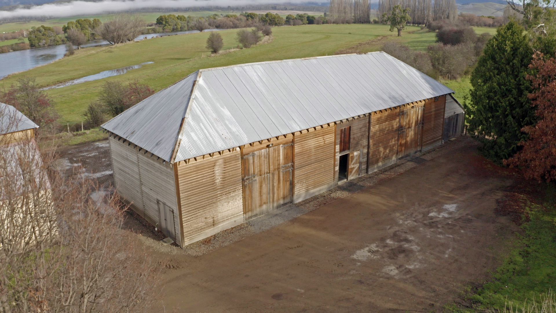 Large wooden barn with a silver roof in a muddy rural field beside a river
