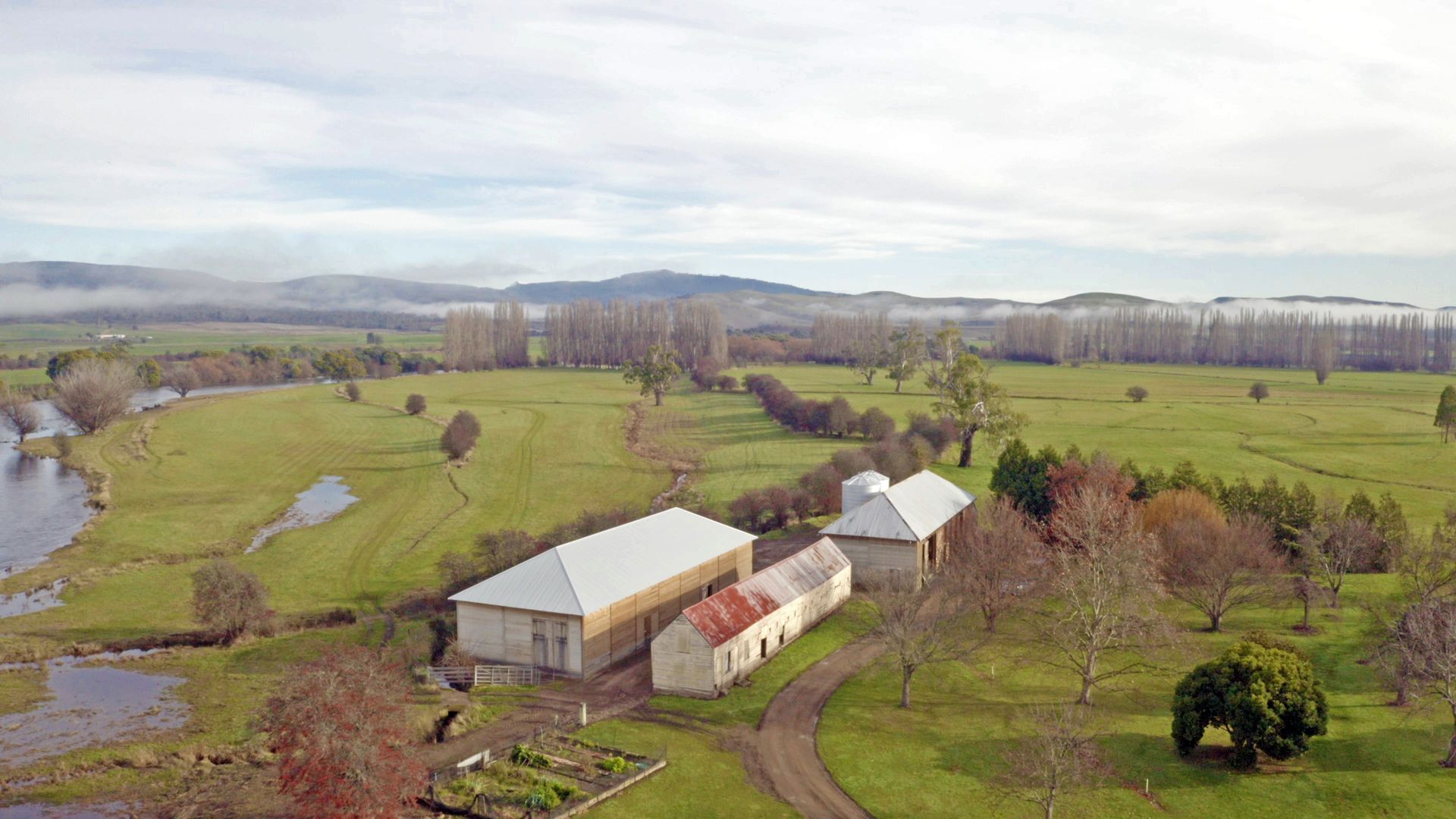 Aerial view of a rural farm with barns, green fields, winding creek, and distant hills under a cloudy sky