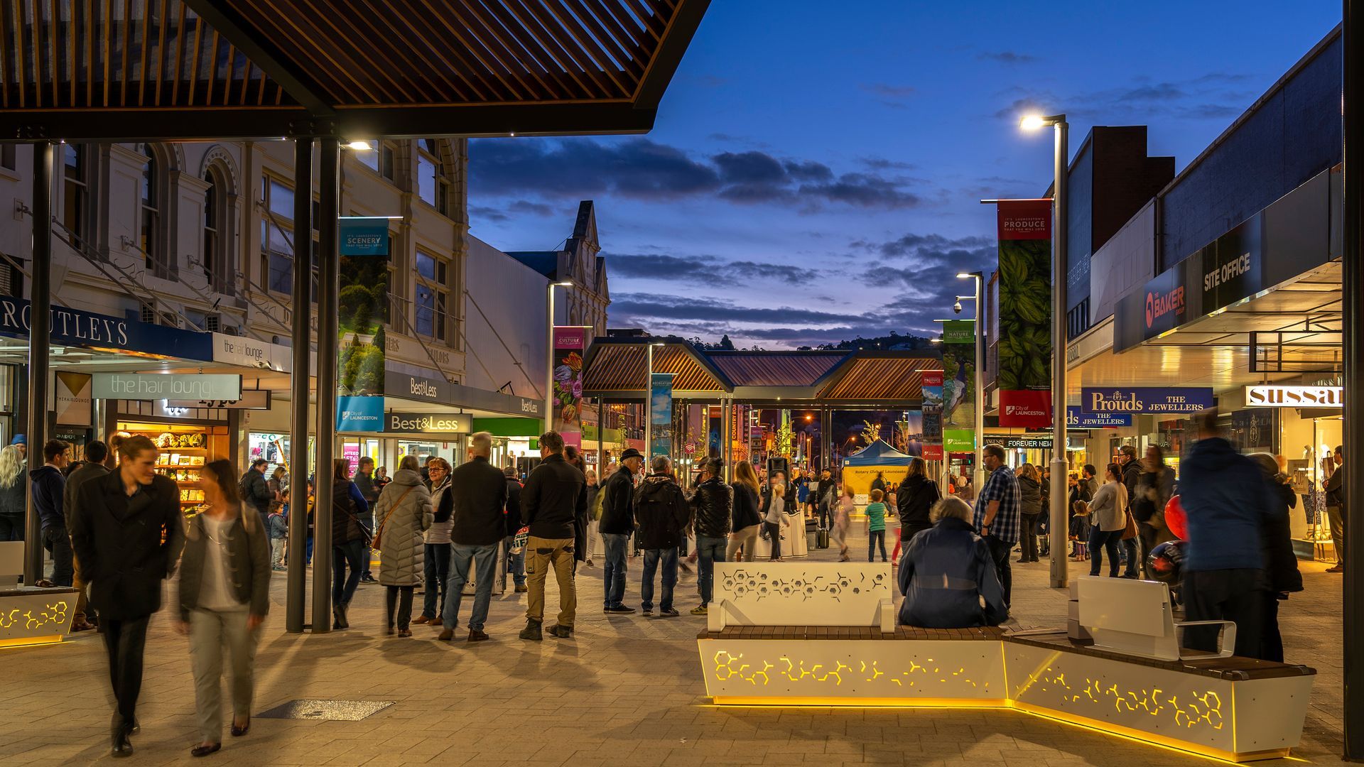 Busy pedestrian shopping street at dusk with lit storefronts, people walking, and a blue-purple sky.