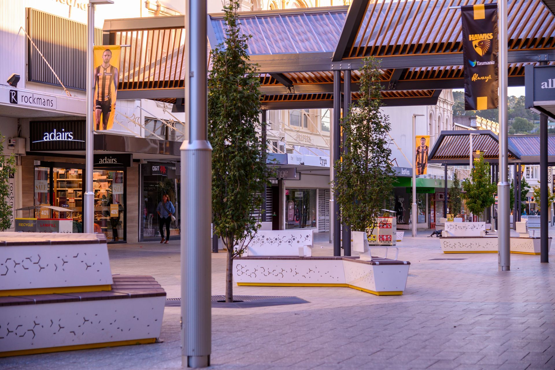 Empty outdoor shopping plaza with benches, trees, and storefronts at dusk