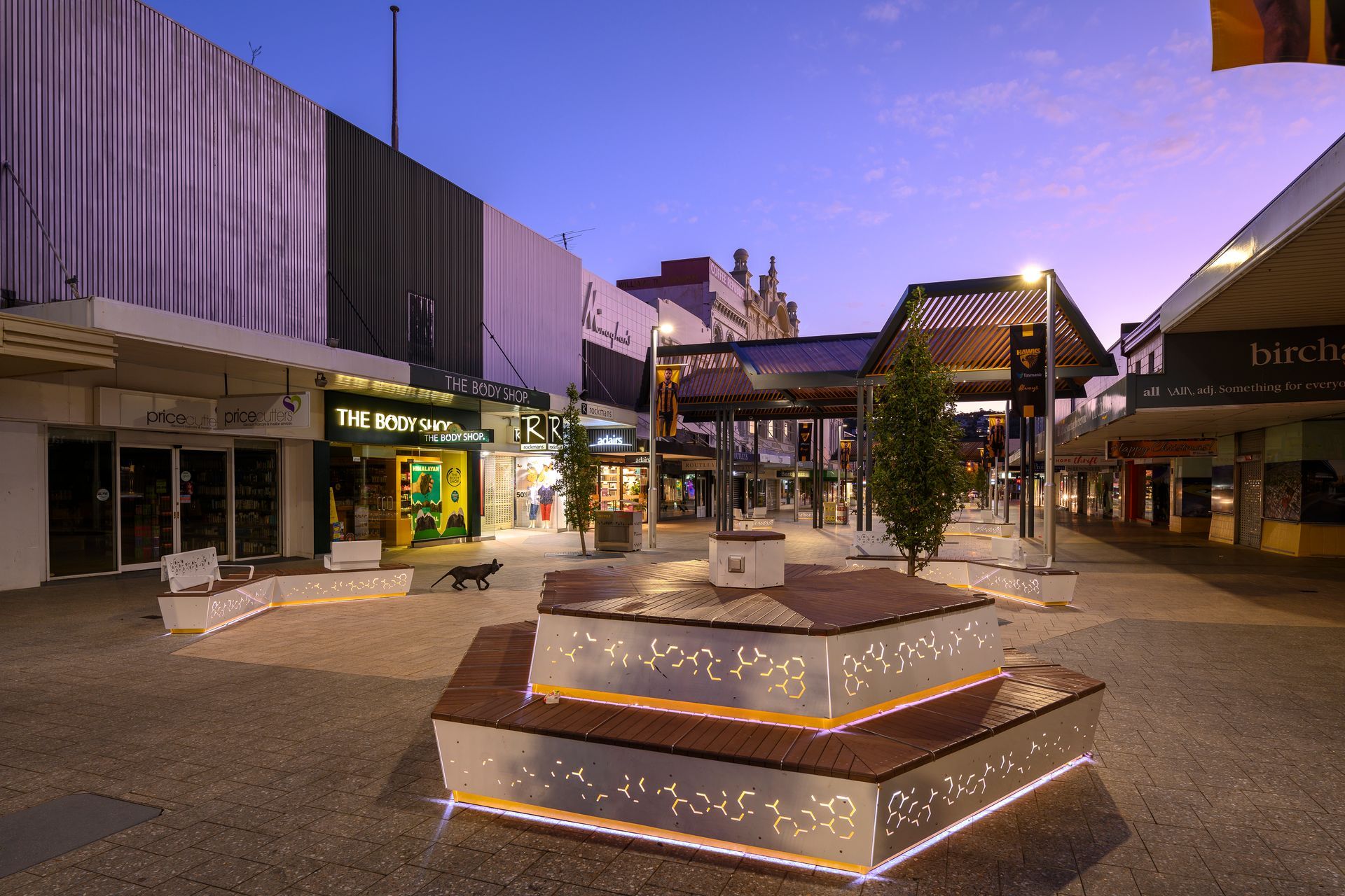 Empty shopping plaza at dusk with lit storefronts and a hexagonal fountain in the center