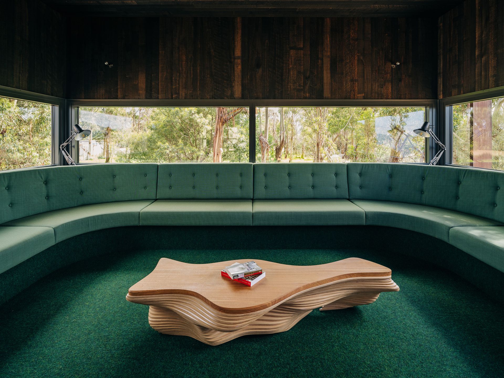 Modern lounge with curved green seating and a sculptural wooden coffee table by large windows.