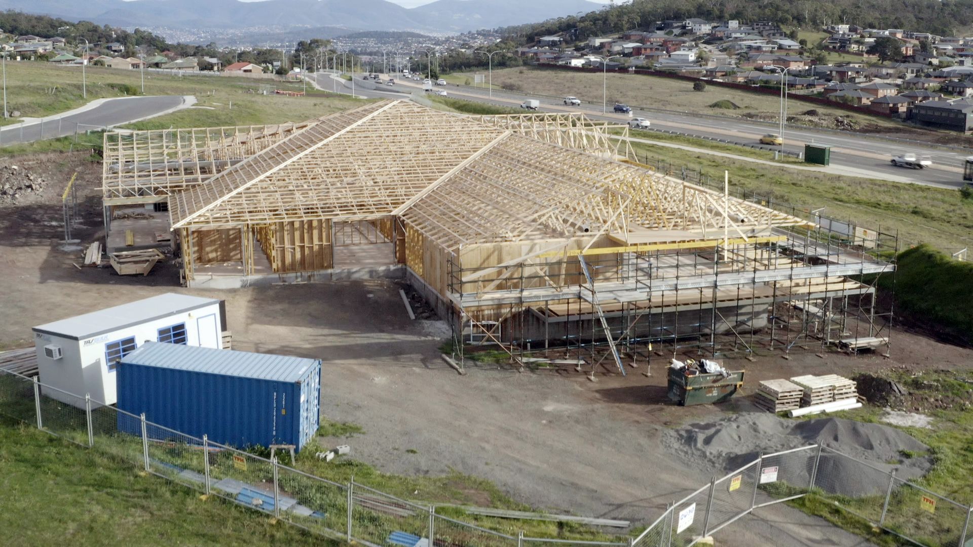 Wooden building frame under construction on a rural site, with hills and roads in the background.