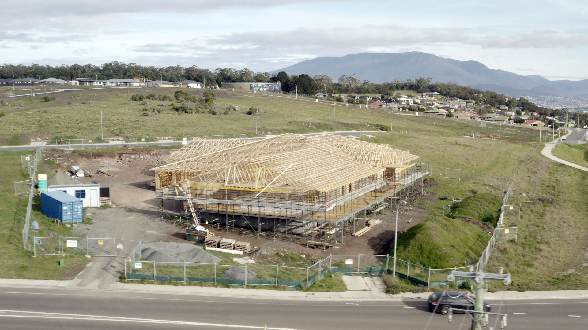 Wood-frame house under construction beside a road in an open field, with mountains in the distance