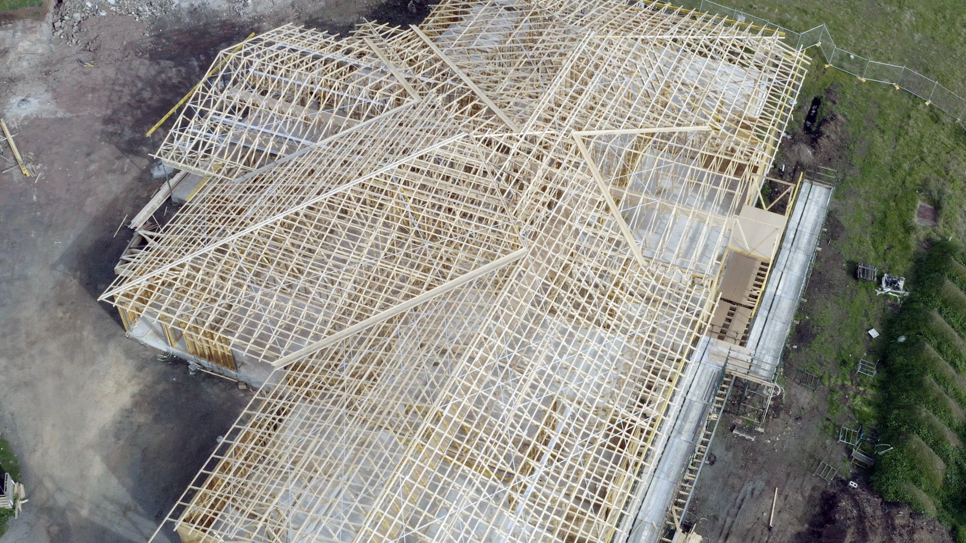 Aerial view of a house under construction with a wooden roof frame and surrounding dirt lot.