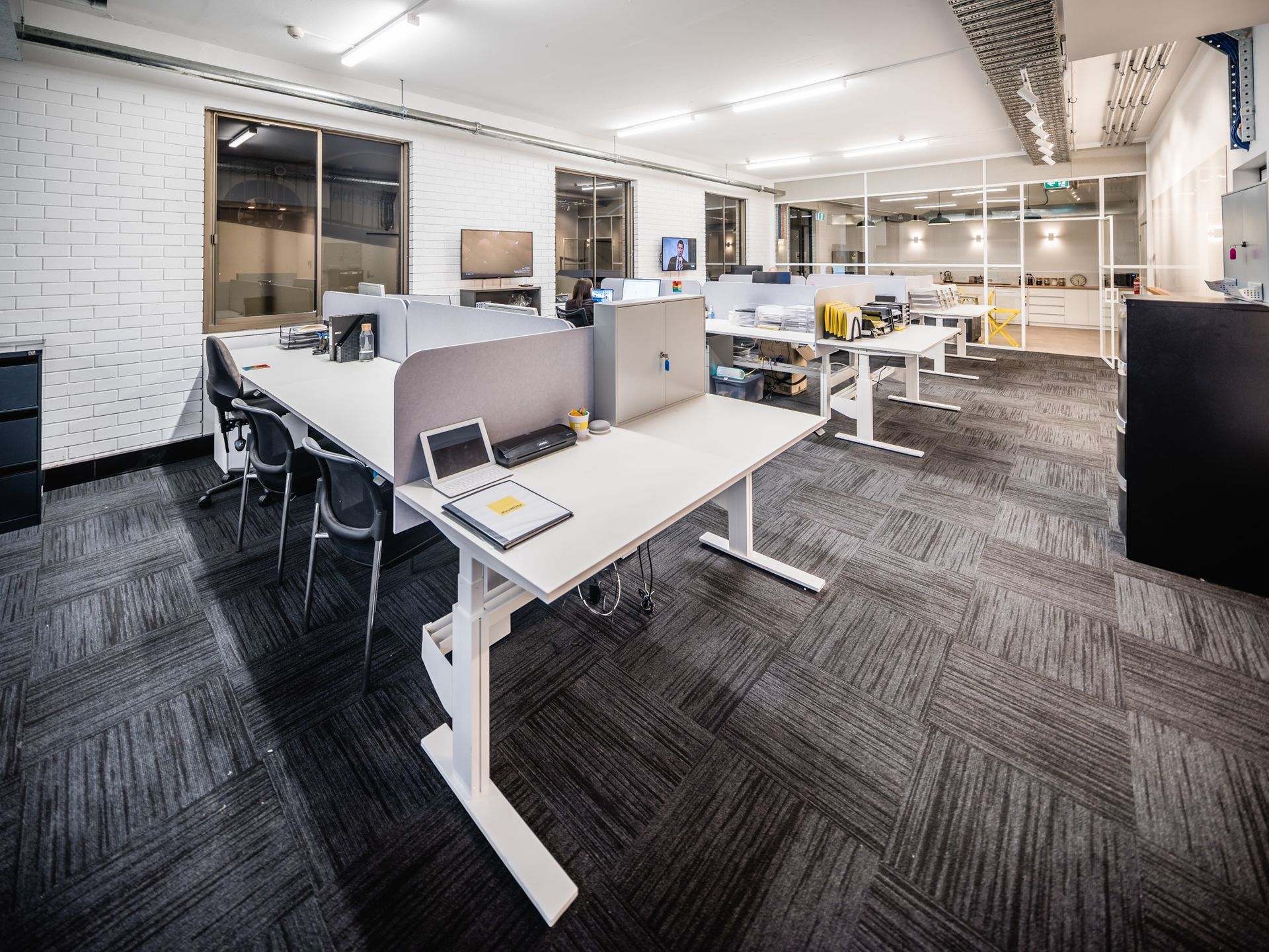 Open-plan office with desks, chairs, computers, and white brick walls under bright lighting