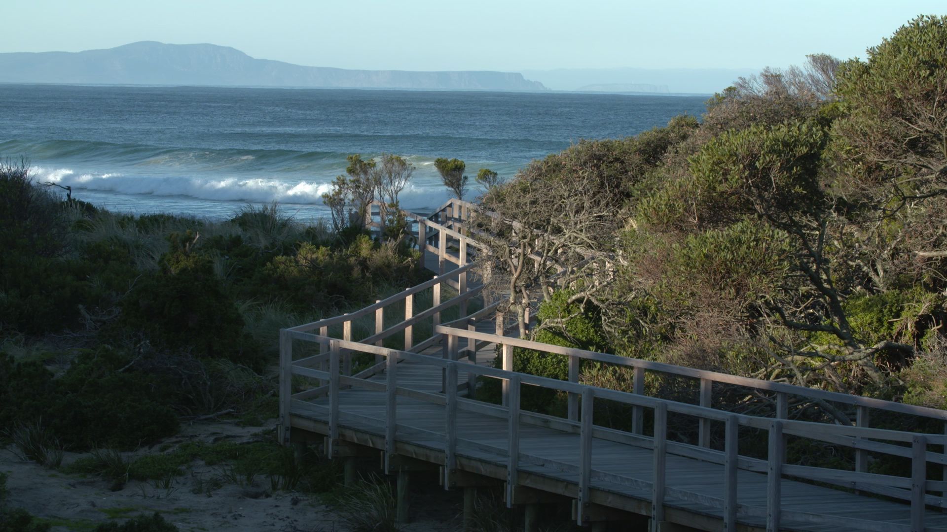 Wooden boardwalk along a coastal path beside waves, dunes, and distant mountains under a clear sky