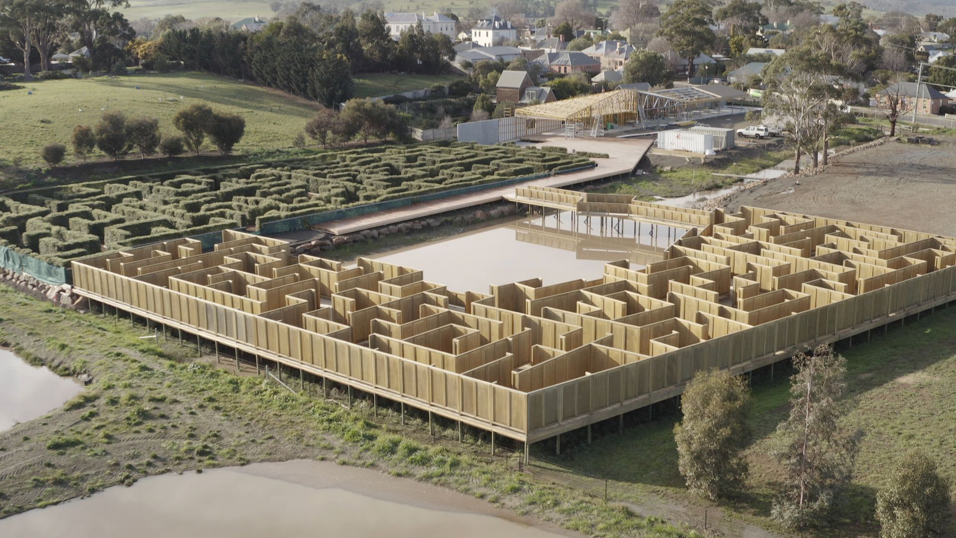 Aerial view of a large wooden maze-like structure in a rural field near a river and houses.