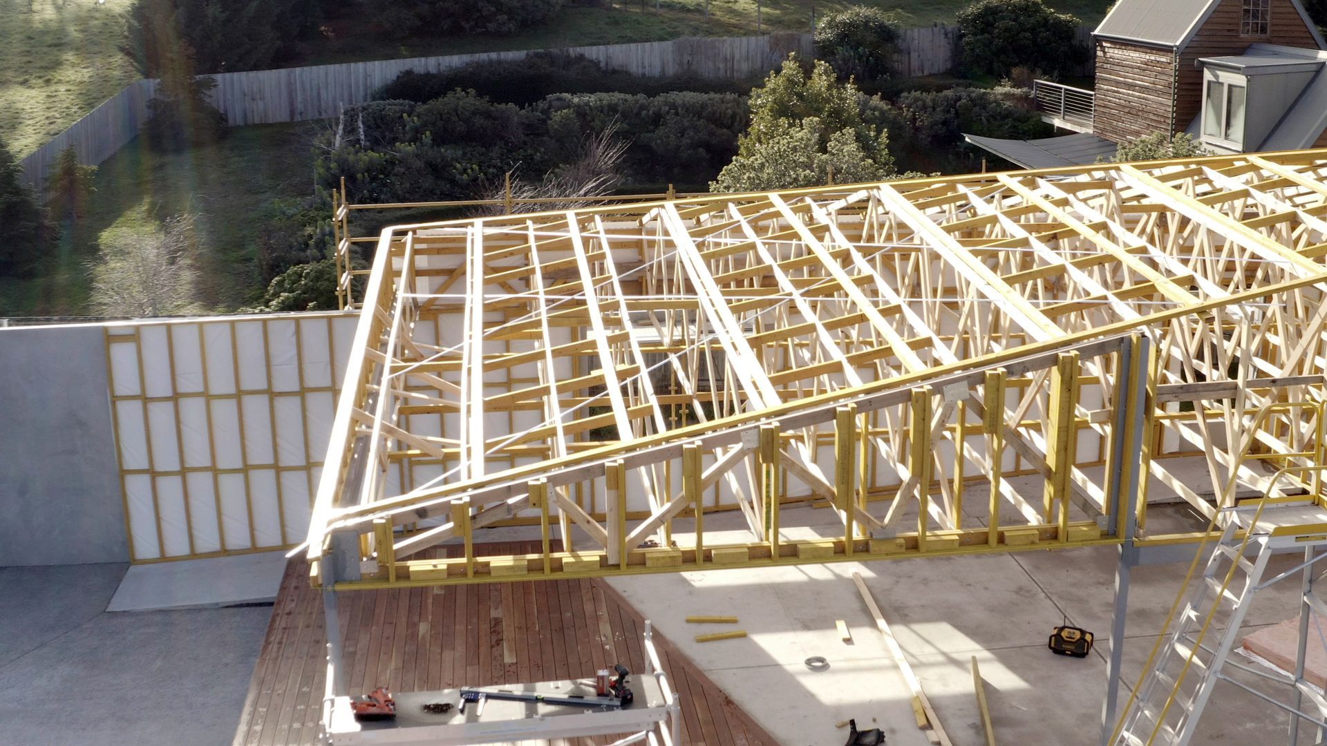 Construction site with a large wooden roof truss frame beside a concrete wall and metal scaffolding