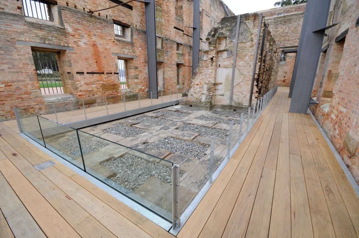 Glass-floored walkway overlooking a ruined brick courtyard with exposed walls and timber decking