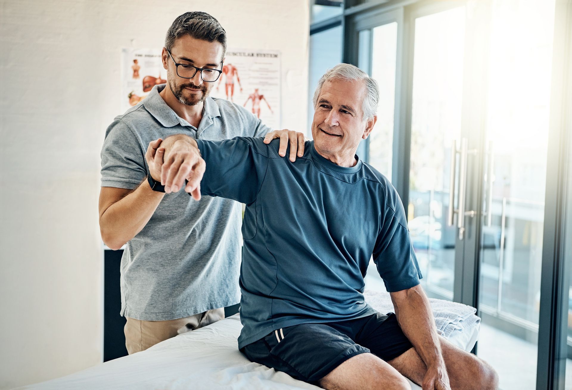 Therapist assisting a smiling seated man during a rehabilitation exercise in a bright clinic