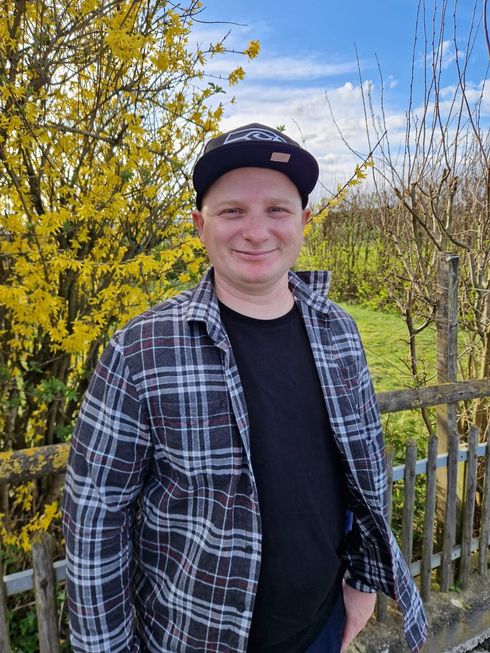 Eine lächelnde Person mit schwarzer Kappe und kariertem Hemd steht im Freien zwischen gelben Blüten und einem Holzzaun.