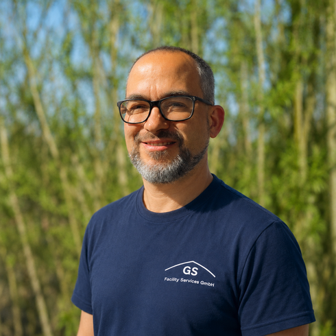 Ein lächelnder Mann mit Brille und schwarzem Polohemd steht im Freien zwischen hohen grünen Pflanzen.