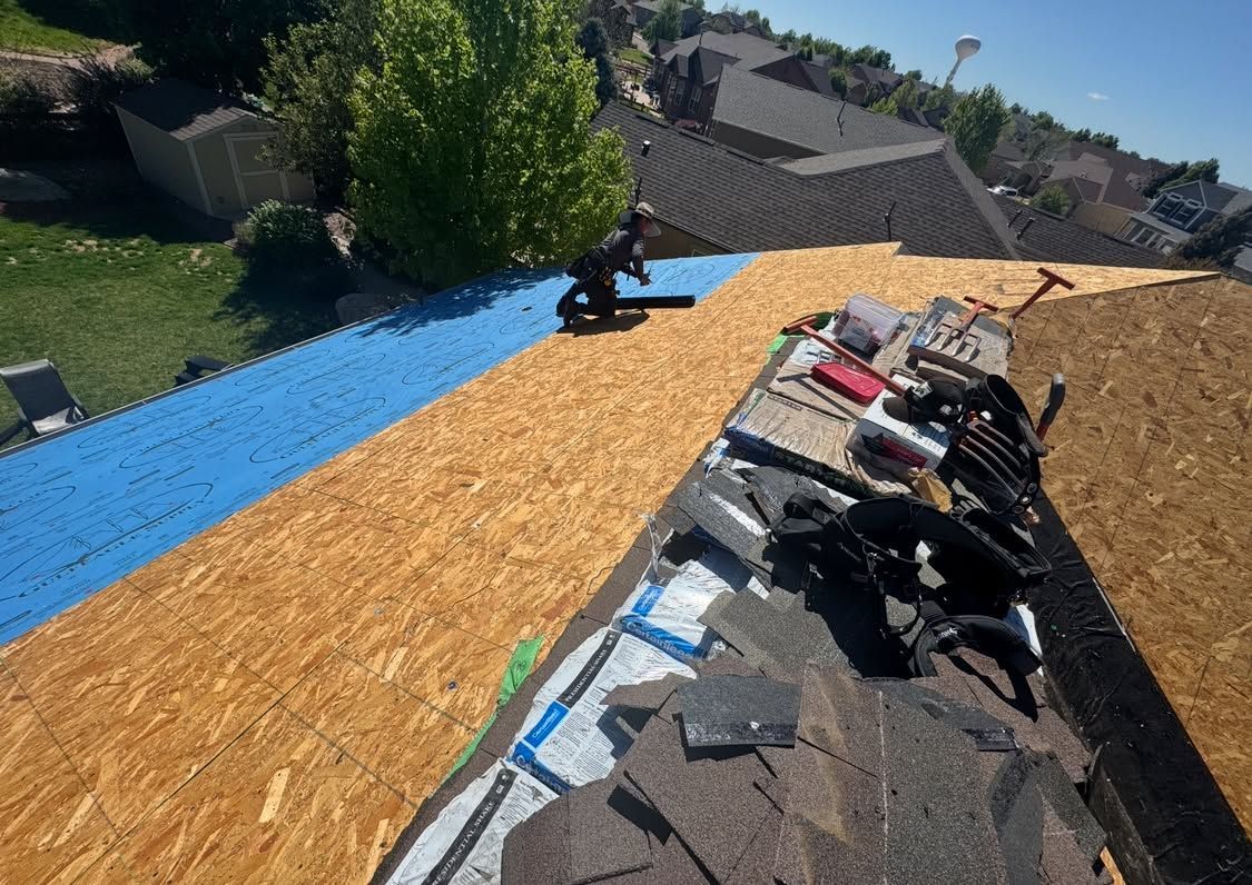 Roof under repair with workers, exposed plywood, blue underlayment, and shingles stacked on the roof.