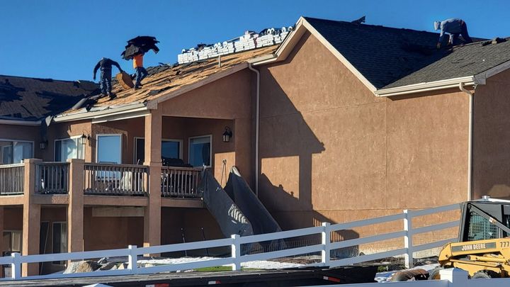 House with roof damage and workers on the roof behind a white fence