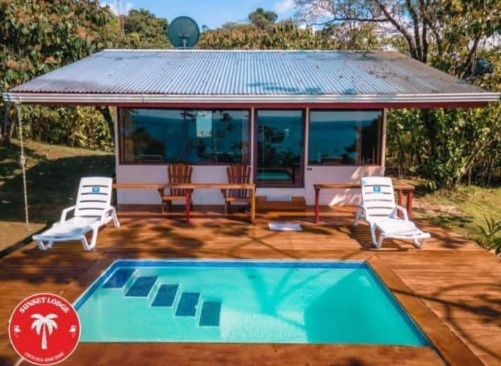 Cottage with a small pool, wooden deck, and lounge chairs. Blue pool, brown wood, and a white building.