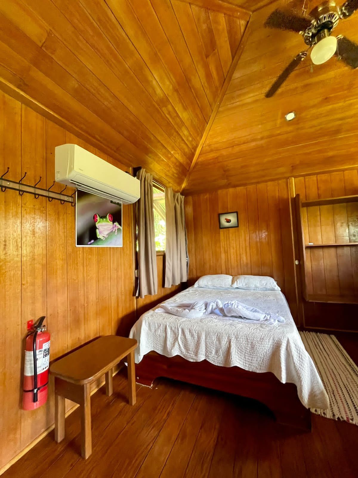 Wooden bedroom with a bed, small table, air conditioner, and fire extinguisher.