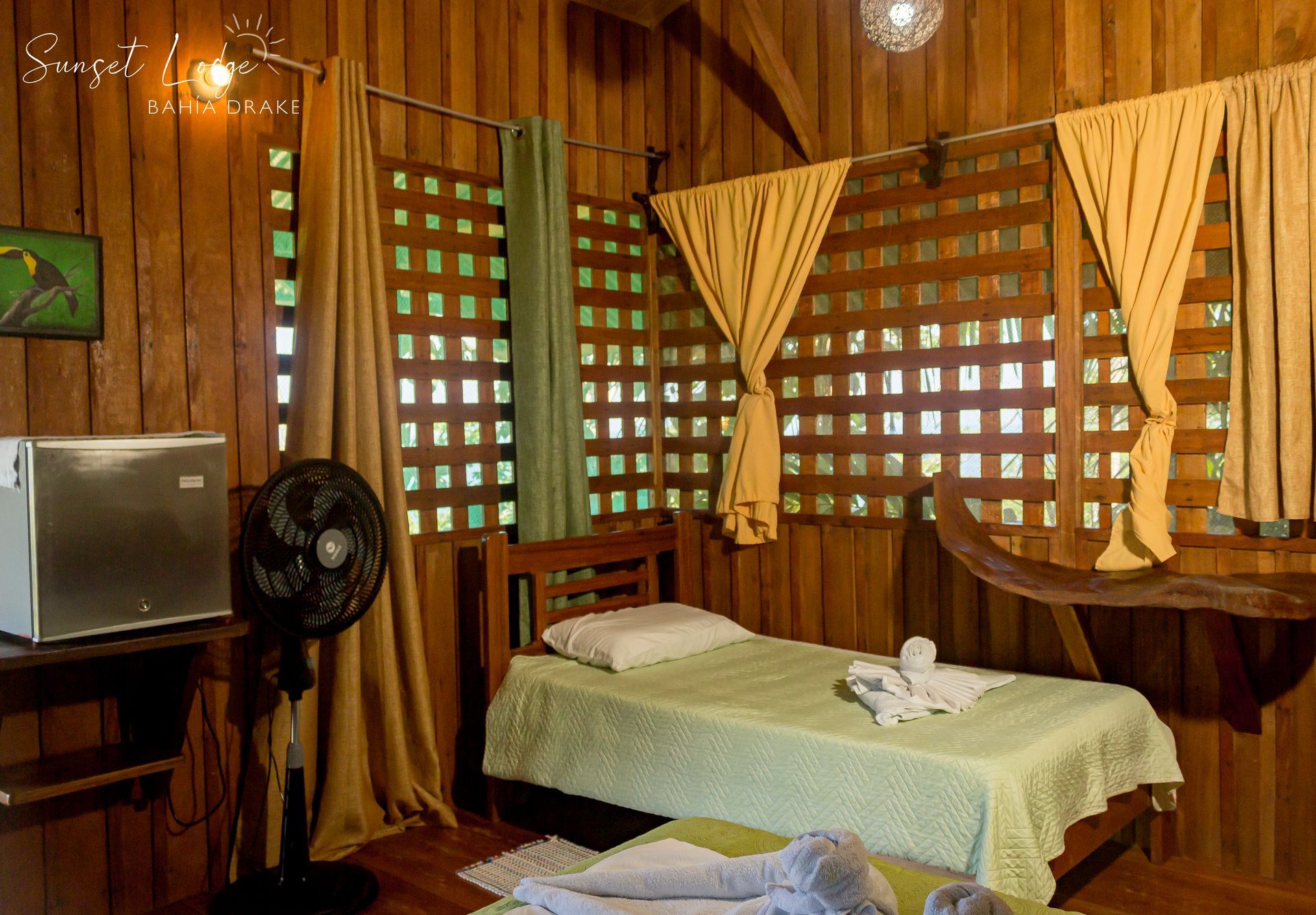 Wooden cabin bedroom with two beds, small refrigerator, fan, and lattice windows, natural light.