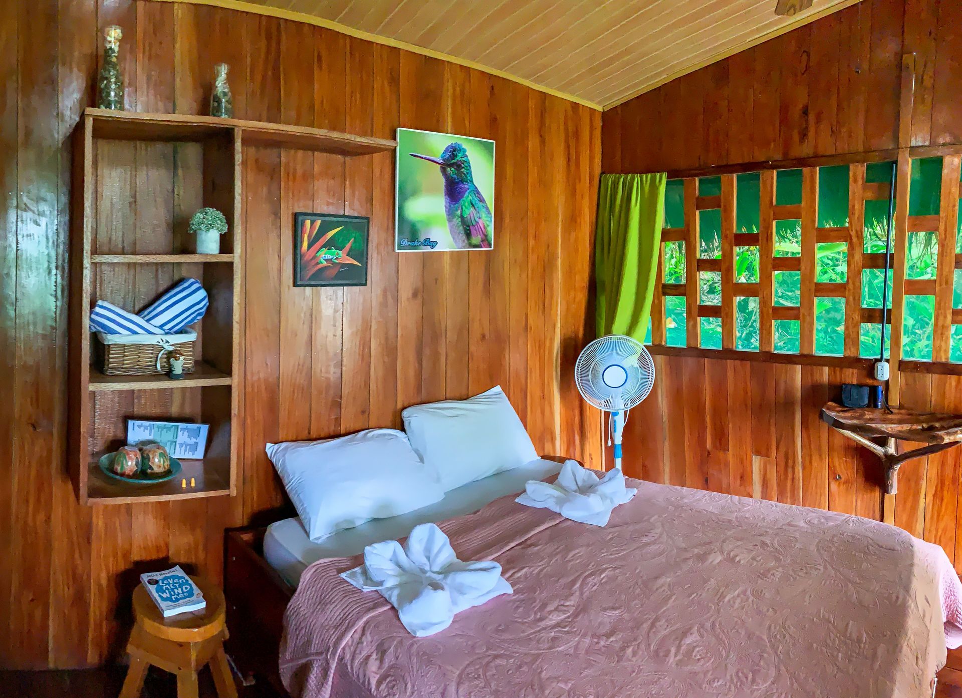 Wooden-walled bedroom with bed, fan, artwork, and open shelf with towels and decorations.