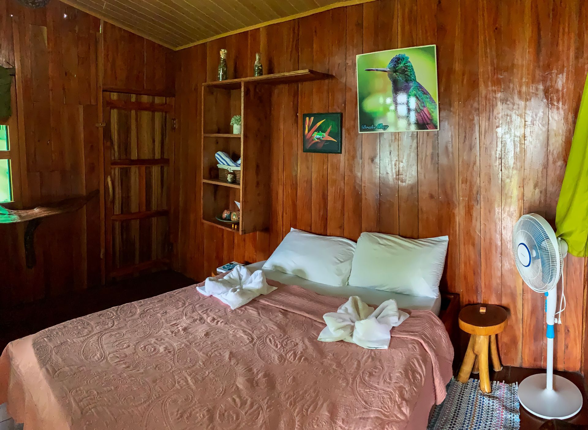 Wooden-paneled bedroom with bed, fan, and hummingbird artwork.