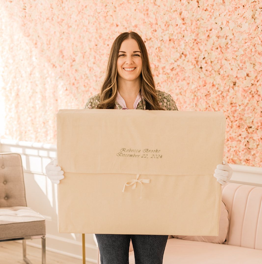 Woman smiles, holding a large beige package with text. Floral background, elegant indoor setting.