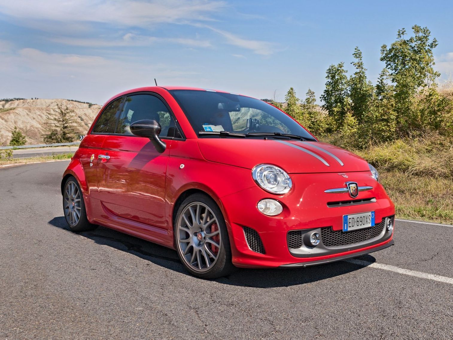 A bright red Abarth 500 sports car parked on an asphalt road in a sunny, rural landscape.