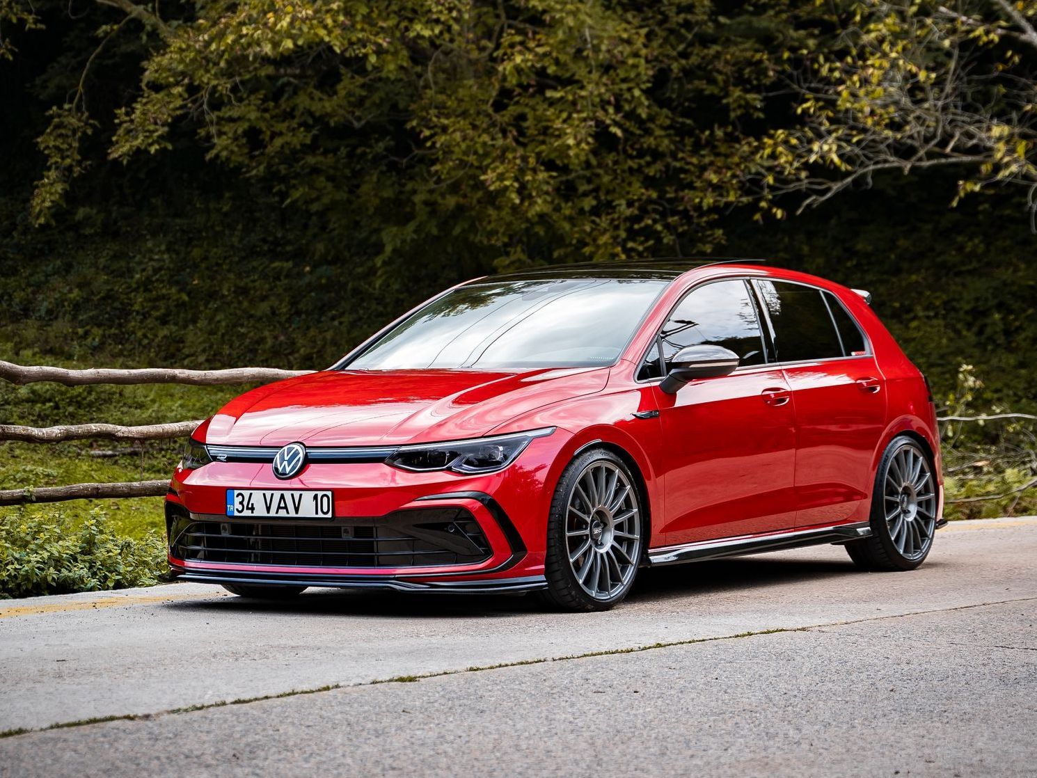 A red Volkswagen Golf hatchback parked on a paved road lined with trees and a wooden fence.