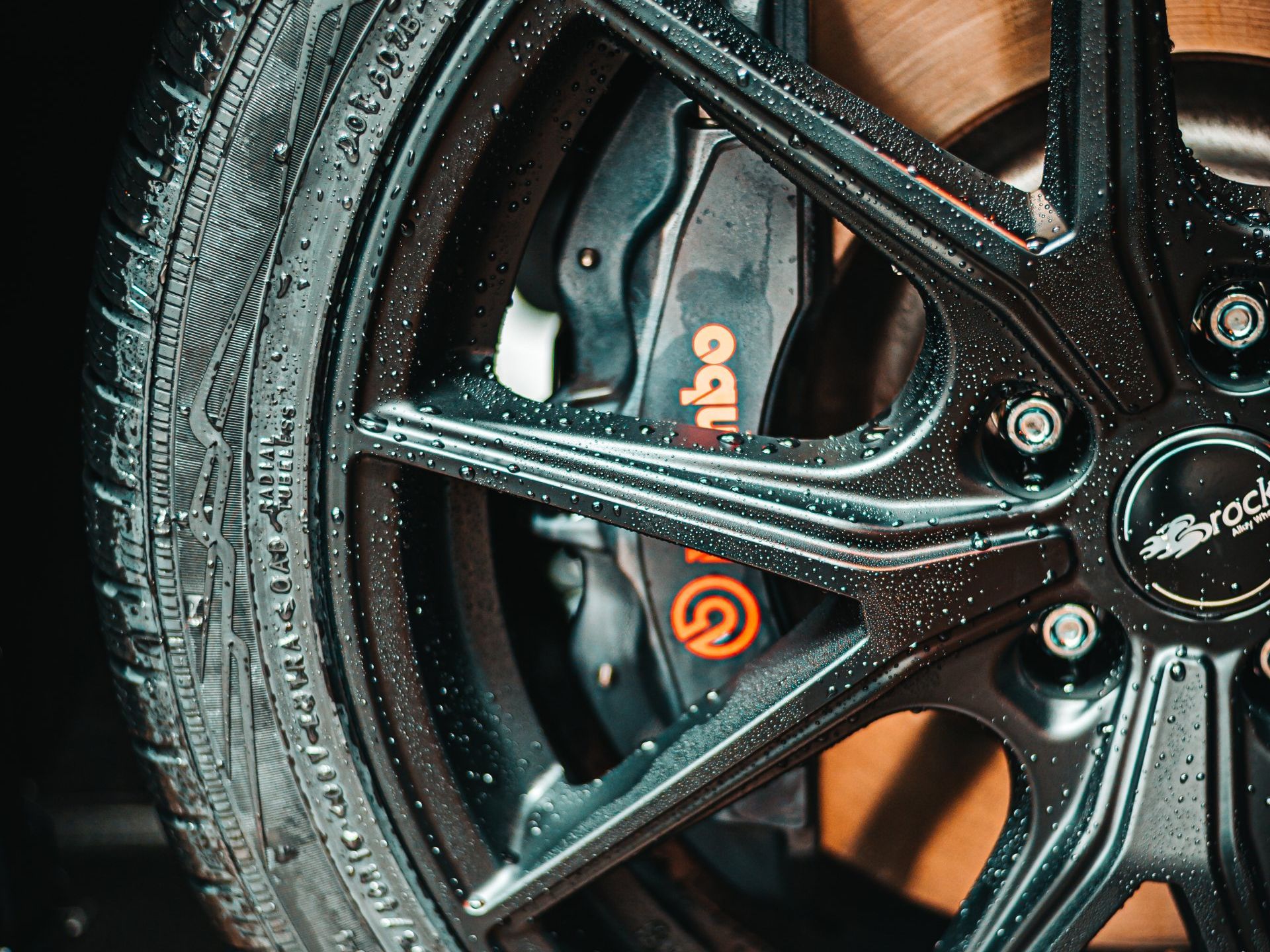 Close-up of a wet black car wheel featuring a prominent orange Brembo brake caliper and a branded center cap.