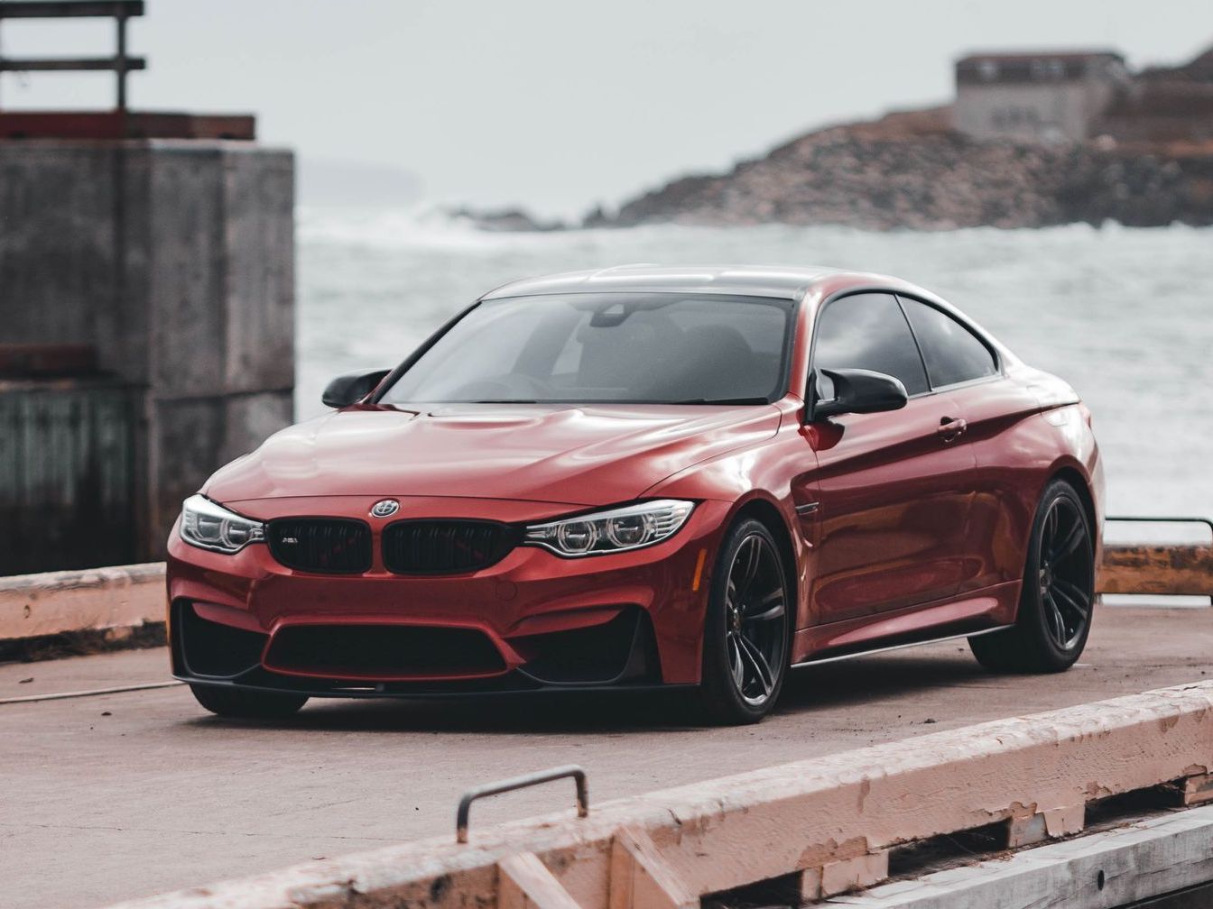A red BMW M4 coupe parked on a concrete pier overlooking a choppy, overcast sea.