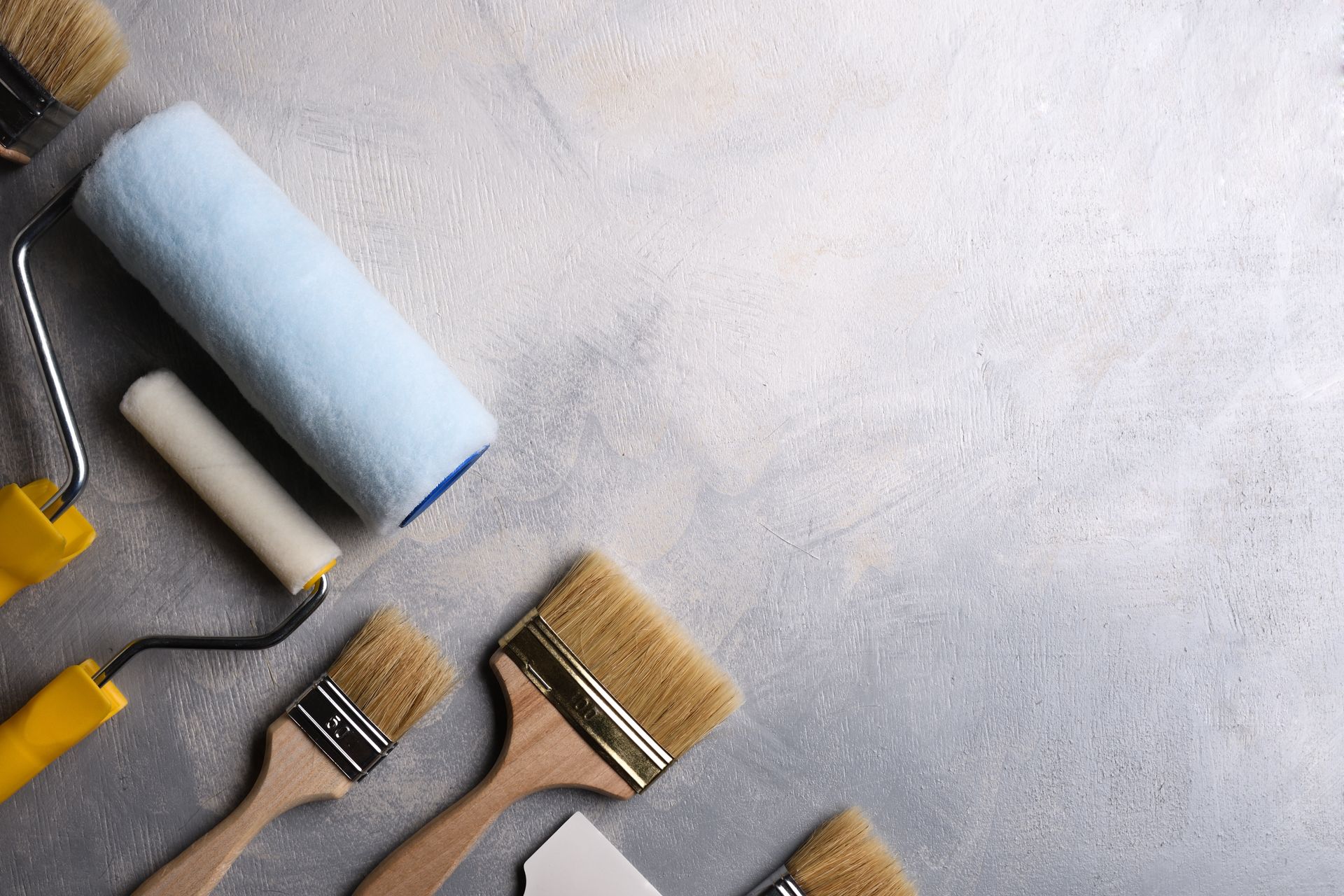 Paint rollers and various paintbrushes arranged on a gray concrete background.