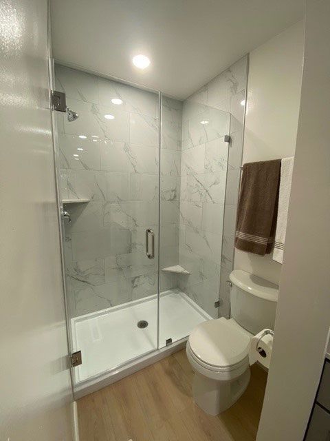 Small modern bathroom with glass shower, white tile, and wood floor; toilet and sink visible.