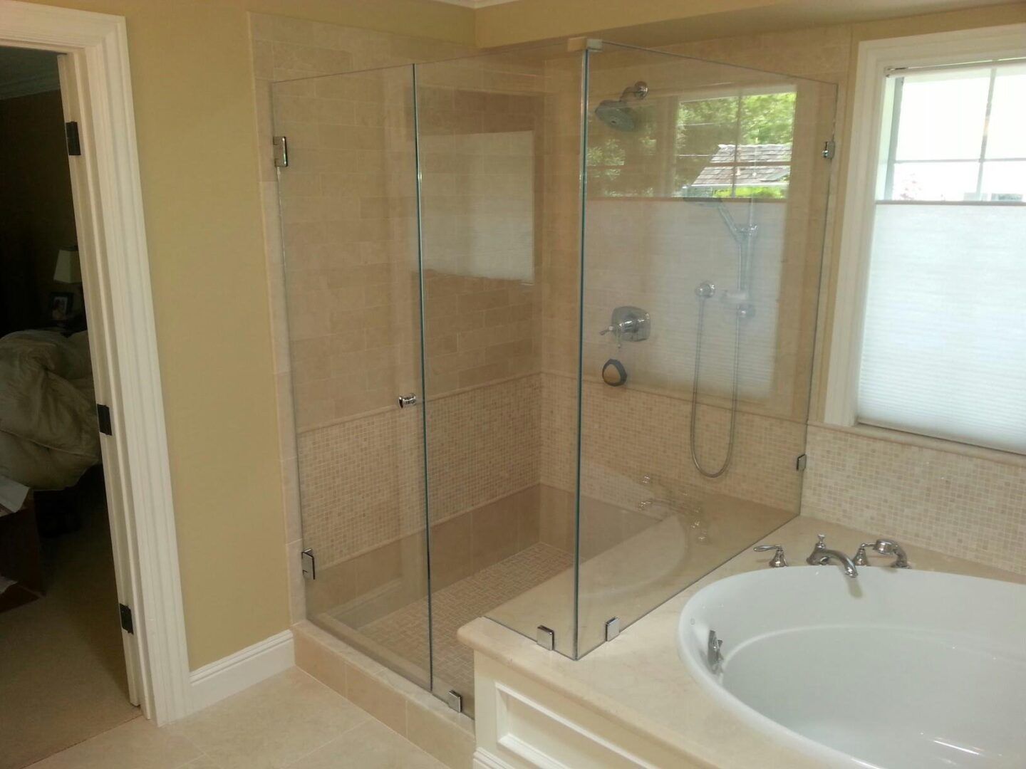 Bathroom with glass-enclosed shower, beige tile, and a white soaking tub beside a window.