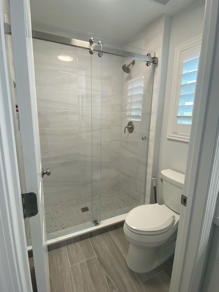 Small bathroom with glass-enclosed shower, white tile walls, and a toilet beside a window.