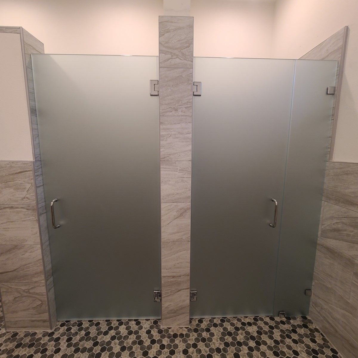 Two frosted glass restroom stall doors with metal handles, separated by a tiled divider.