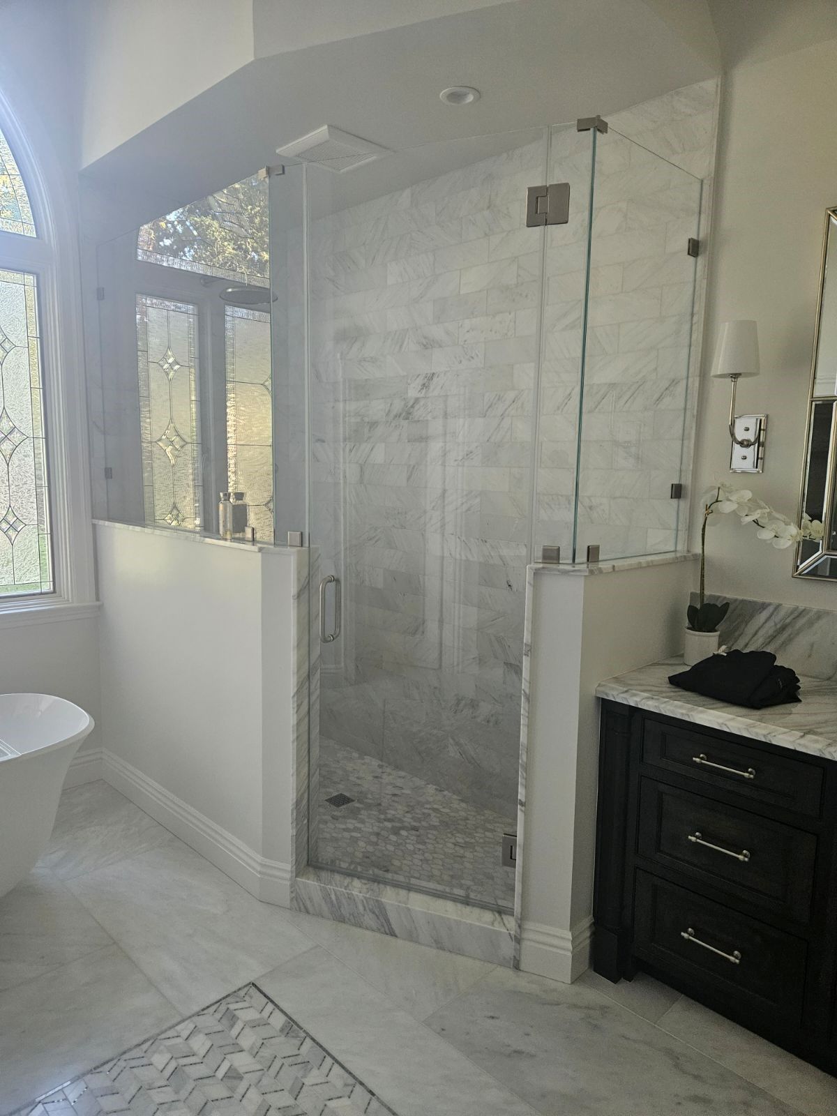 Modern bathroom with glass shower, white tile, large window, and dark vanity.