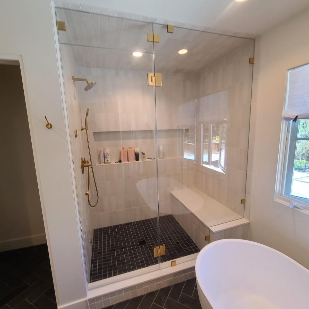 Modern glass walk-in shower with black tile floor, built-in shelf, and white freestanding tub nearby