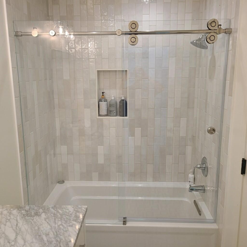 White tiled bathroom with a bathtub, glass shower wall, and built-in shelf of toiletries