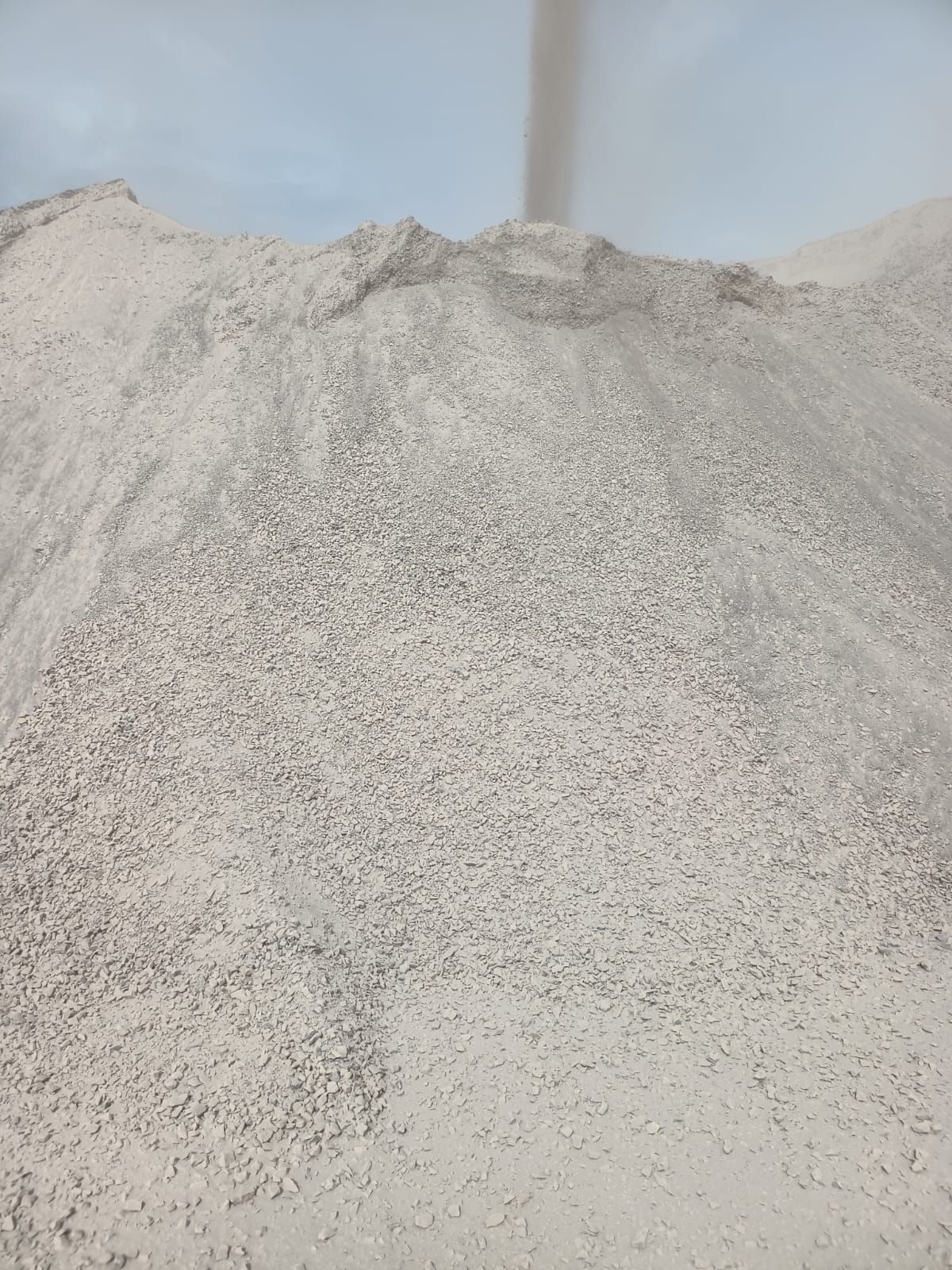 Montón de material granular gris, posiblemente arena o grava, bajo un cielo nublado.