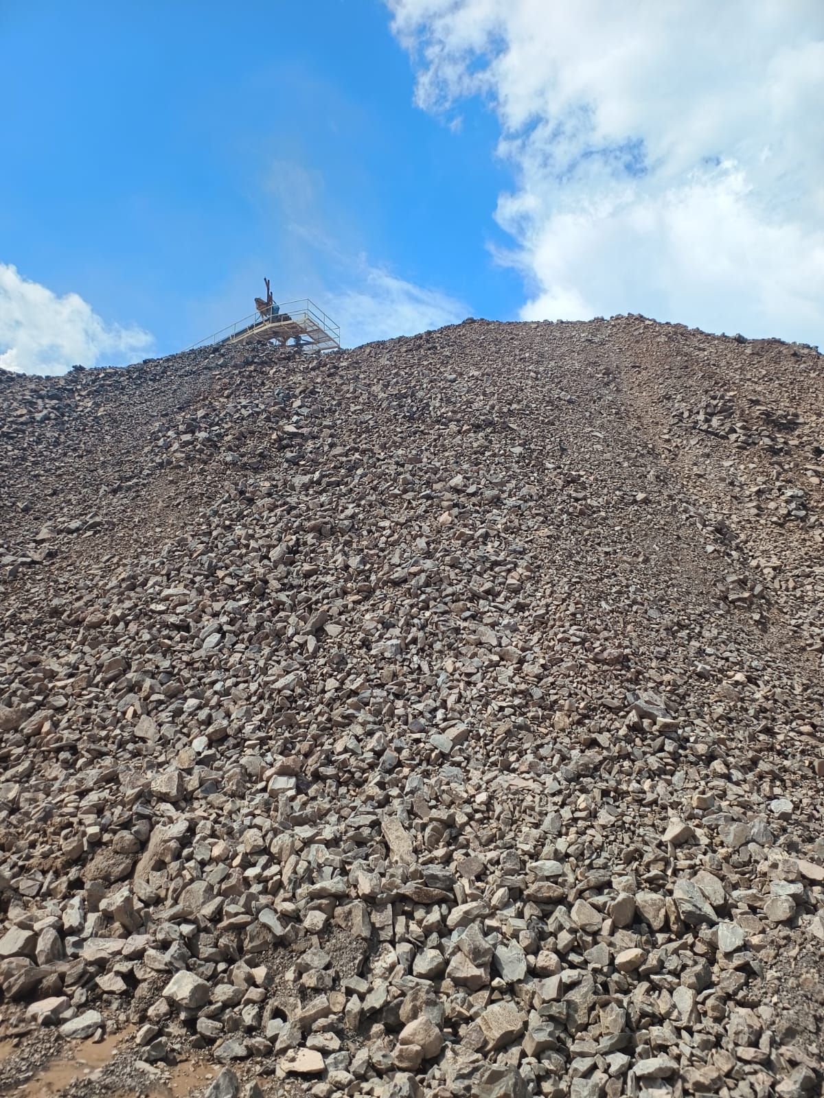Gran montón de material oscuro y rocoso bajo un cielo azul con una máquina trabajando en la cima.