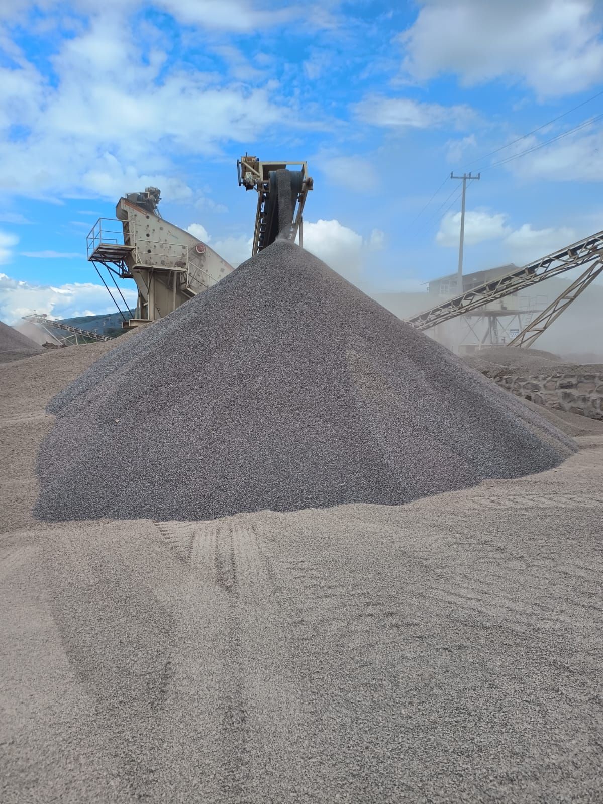 Montón de grava gris en un entorno industrial, con maquinaria al fondo y un cielo azul nublado.