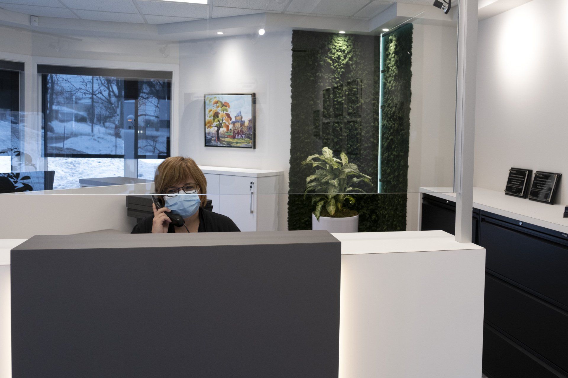 Une femme portant un masque répond au téléphone à son bureau, devant un mur vert et une fenêtre.