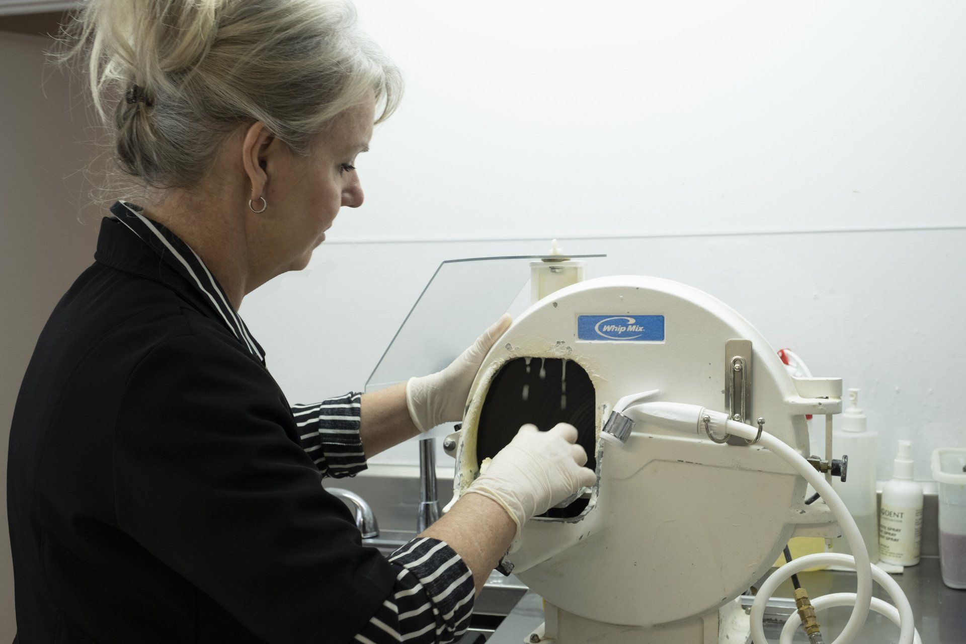 Femme utilisant une machine de laboratoire dentaire, portant des gants, dans un décor aux murs blancs.