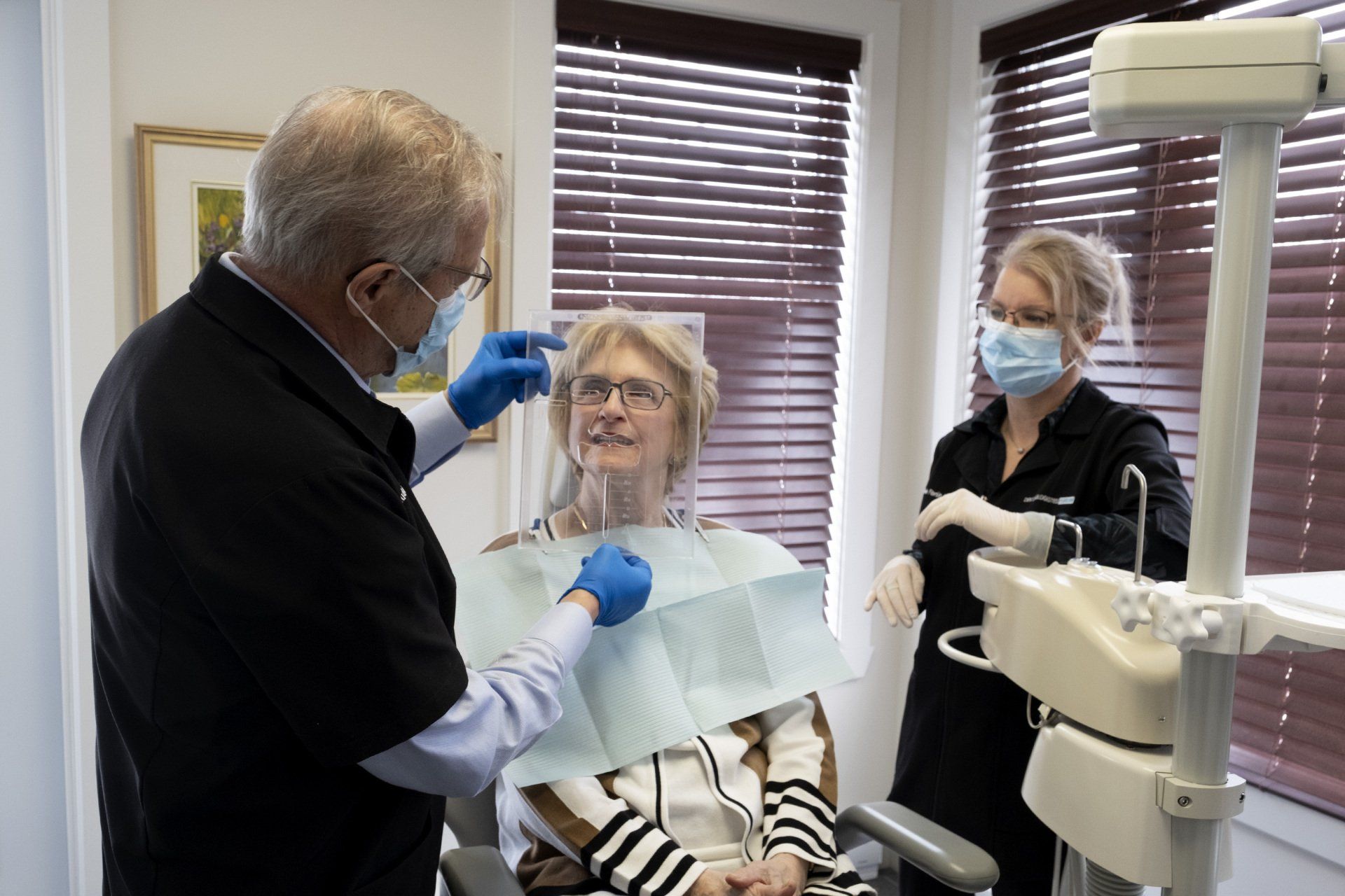 Un dentiste ajuste une visière de protection sur un patient assis sur un fauteuil dentaire, avec un assistant à proximité.