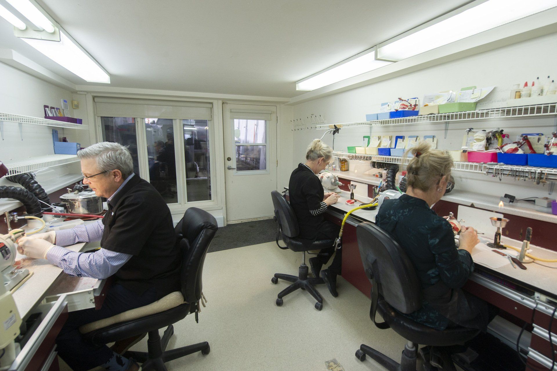 Trois personnes travaillent à des établis dans un atelier bien éclairé. Assises, elles sont concentrées sur leurs tâches.