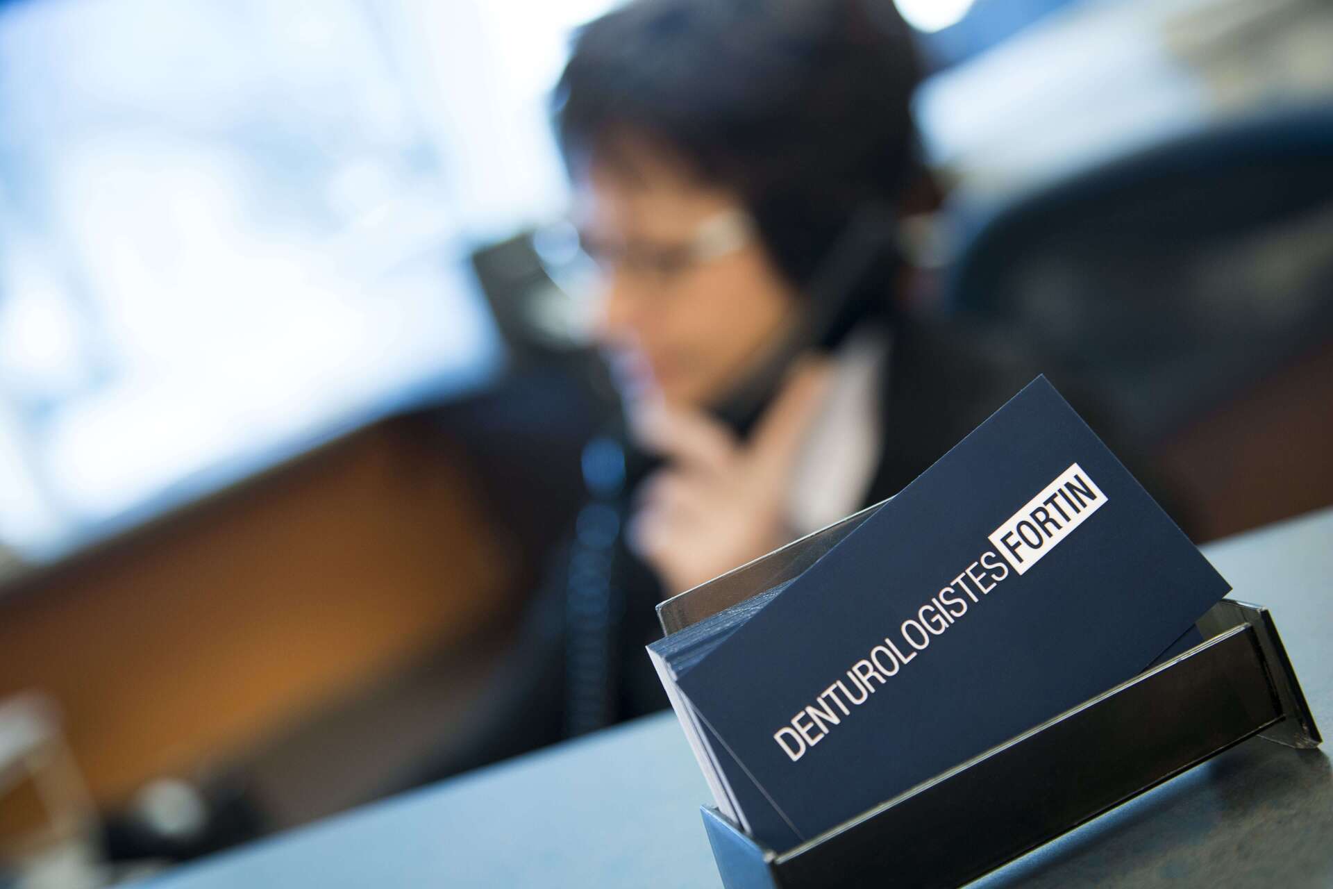 Cartes de visite, « DENTUROLOGISTES FORTIN », sur un comptoir, personne au téléphone en arrière-plan flou.